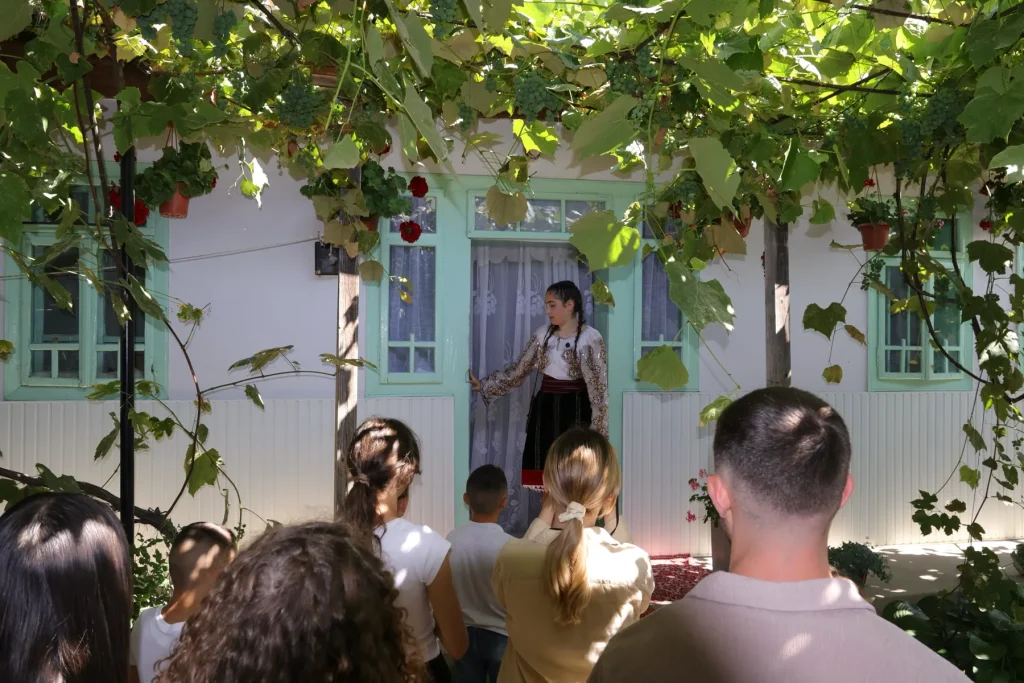 Gazdă în costum popular povestind vizitatorilor sub bolta de viță, la Casa Cojocarului Opincă