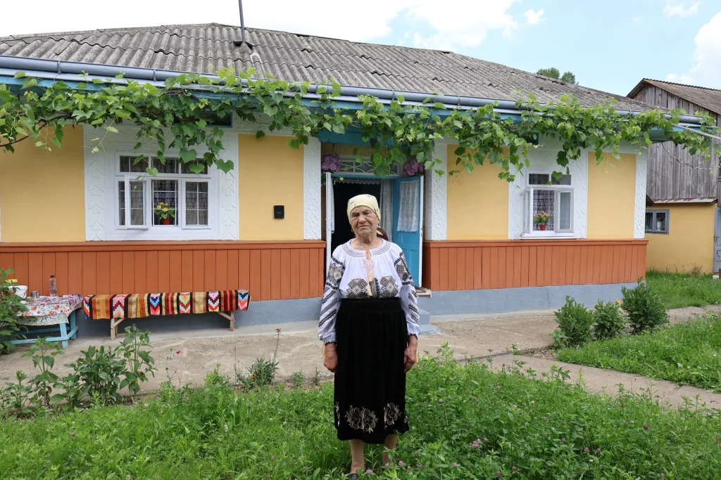 Gospodină în costum popular în fața Casei Veronica, cu scoarțe țesute întinse la soare