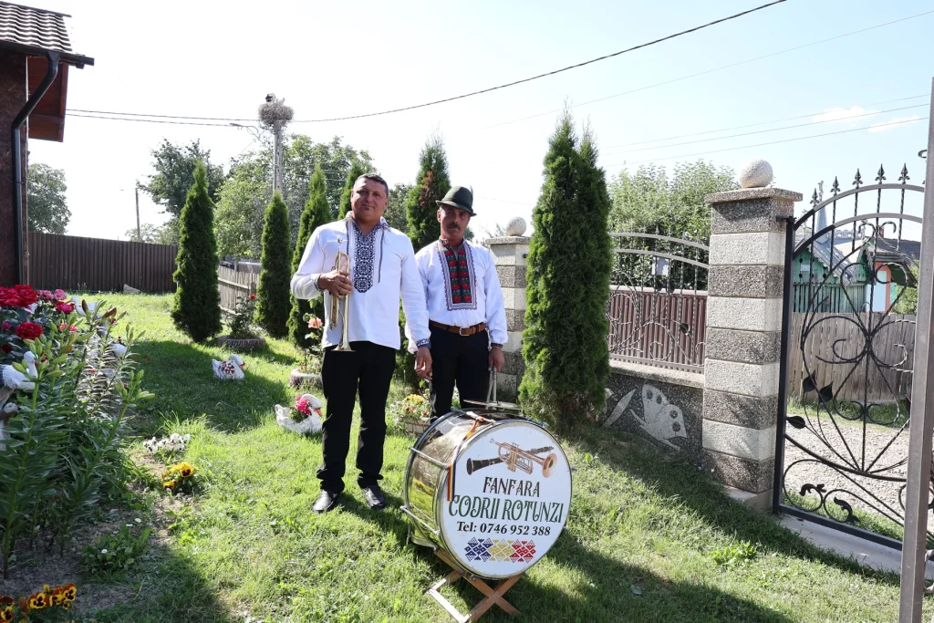 Doi membri ai Fanfarei Codrii Rotunzii în costume populare, lângă toba fanfarei